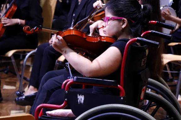 Joven toca violín en silla de ruedas en el escenario junto a una orquesta